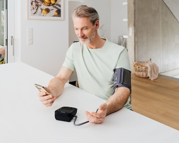 Mann sitzt am Tisch, misst Blutdruck mit Oberarmmessgerät und blickt lächelnd auf sein Smartphone.