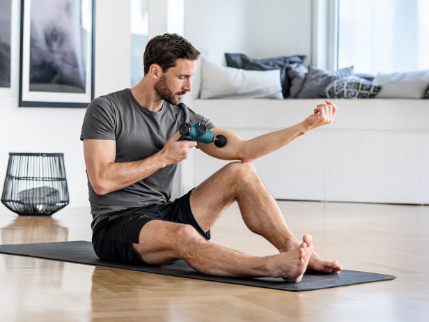 Utilisation du pistolet de massage MG 99 de Beurer Homme assis sur un tapis de sport en tenue de sport et appliquant le pistolet de massage MG 99 de Beurer