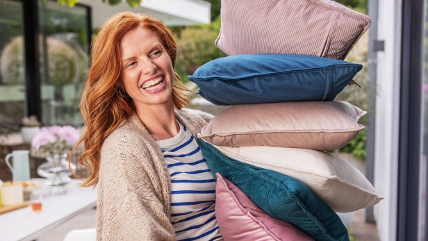 Rothaarige Frau lächelt in die Kamera und hält alle 6 Farben des Heizkissens Heaty von Beurer in den Armen