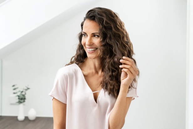 Frau mit langen, gewellten Haaren lächelt und schaut zur Seite in einem hellen, modernen Raum