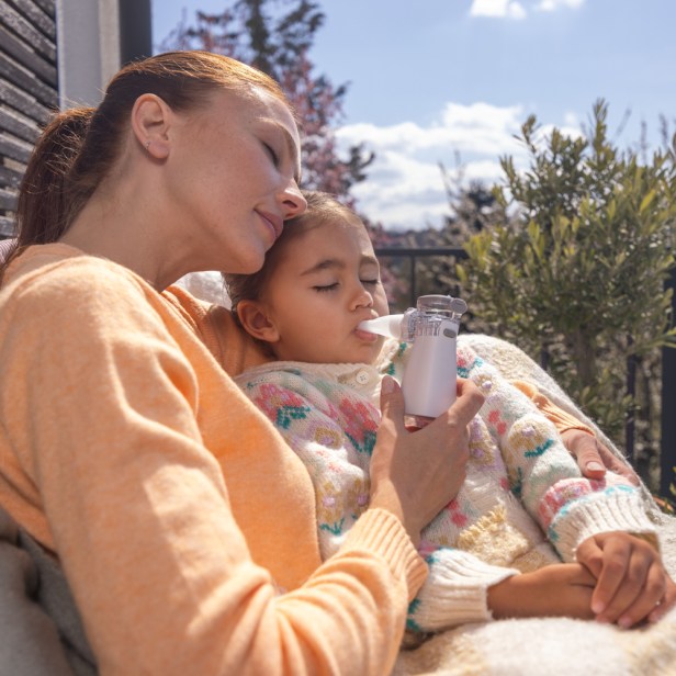 Eine erwachsene Person hält ein Kind im Arm und nutzt dabei ein kleines medizinisches Inhalationsgerät im Freien auf einer sonnigen Terrasse.