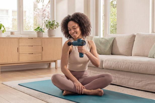 A woman is massaging her shoulder with a Beurer massage gun at home, on a yoga mat.