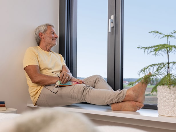 Wohlfühlatmosphäre zu Hause: Ein lächelnder Mann sitzt mit einem Buch entspannt auf der Fensterbank neben einer grünen Zimmerpflanze.
