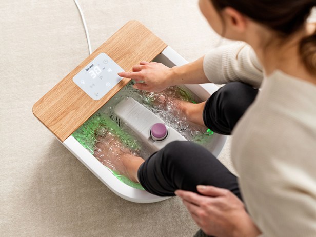 Frau verwendet Beurer MP 65 Fußsprudelbad Entspannendes Fußbad: Eine Frau bedient das Touch-Display ihres Beurer Sprudelbads mit Holzdekor und grün beleuchtetem Wasser.