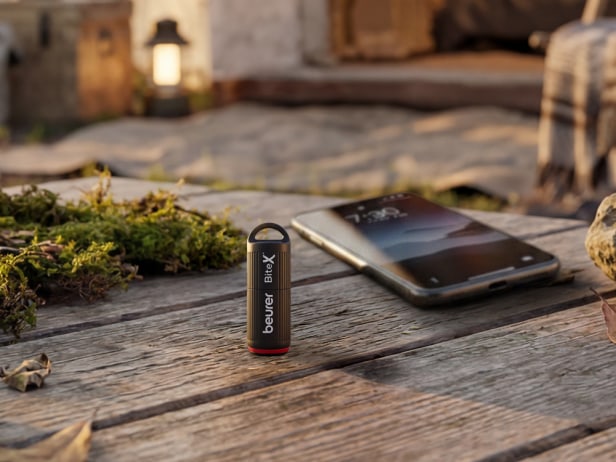The Beurer BitexX Go is sitting next to a smartphone on a wooden table outside.