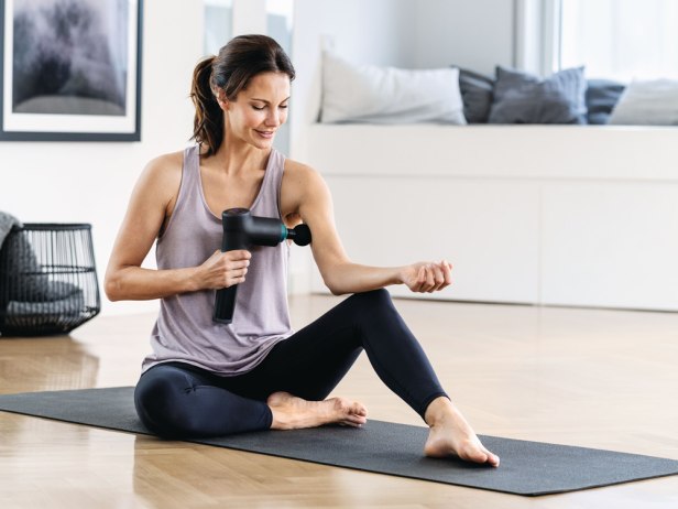 Une femme se masse l'épaule avec un pistolet de massage Beurer, chez elle, sur un tapis de yoga.