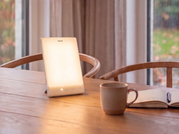 La lampe à lumière du jour Beurer TL se trouve dans la salle à manger Une lampe TL 45 à lumière du jour est posée sur une table à manger, à côté d'une tasse de café et d'un livre ouvert
