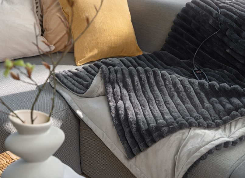 Graue Beurer Heizdecke Stockholm, schön platziert auf einem hellgrauen Sofa mit gelben Sofakissen