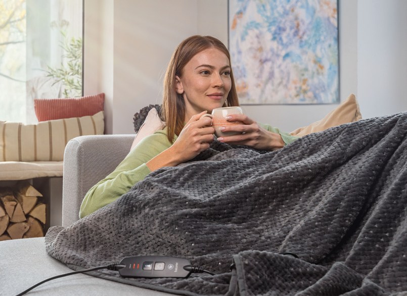 Frau mit roten Haaren liegt auf einem Sofa unter der Beurer Heizdecke HD 65 und trinkt Tee