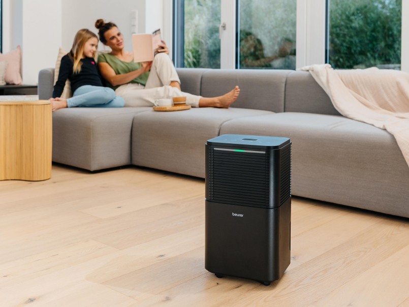 Der Luftentfeuchter steht in einem Wohnzimmer auf dem Boden, im Hintergrund sitzen eine Frau und ein Mädchen auf dem Sofa