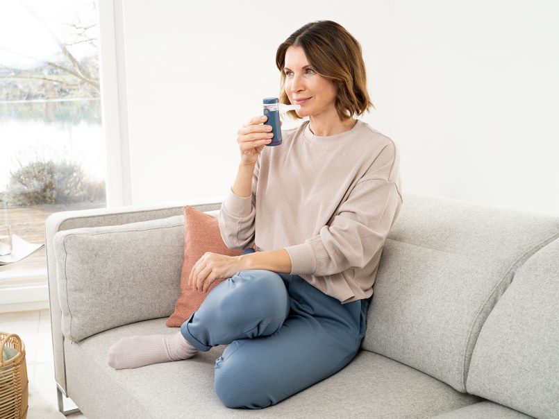 Eine Frau sitzt entspannt auf einem Sofa im hellen Wohnzimmer und nutzt einen Inhalator für ihre Atemgesundheit.