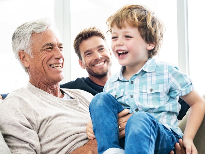 Drei Generationen einer Familie – Großvater, Vater und Enkel – lachen herzlich zusammen auf dem Sofa im Wohnzimmer.