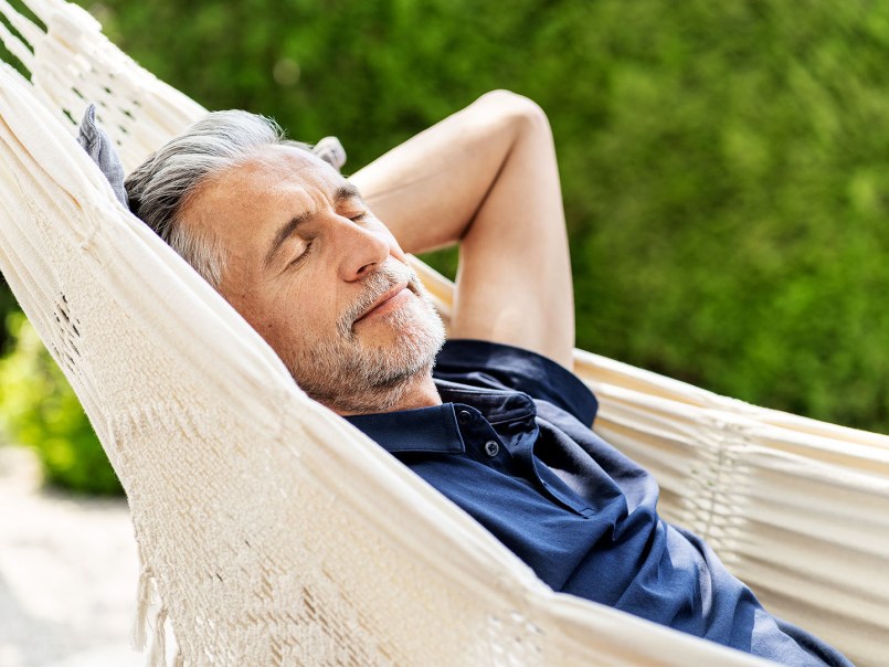 Mann entspannt in Hängematte im Garten, hat die Arme über dem Kopf verschränkt und die Augen geschlossen und wirkt zufrieden