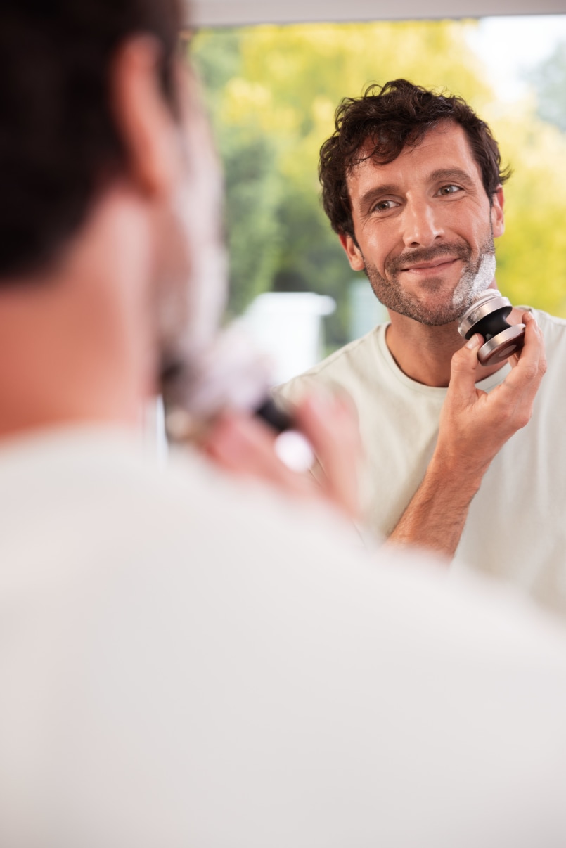 Mann reinigt sein Gesicht mit der Beurer FC 59 Dual Gesichtsbürste und lacht sich dabei im Spiegel im Badezimmer an