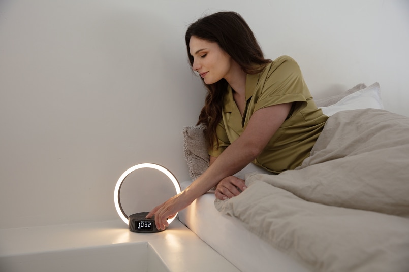 Frau liegt im Bett und ist zu ihrem Nachttisch gebeugt. Auf diesem steht das Wl 65, auf diesem lädt ein Handy. Die Frau berührt ganz sanft das Handy. Das WL 65 ist an und leuchtete.  