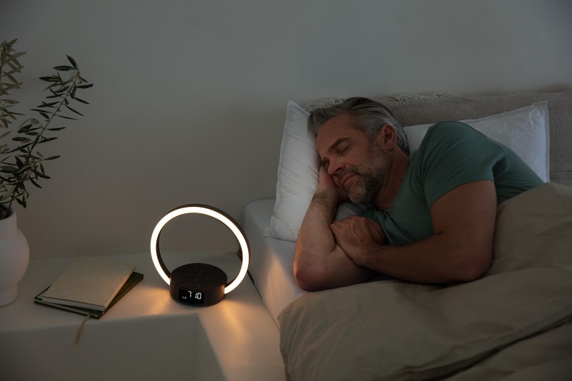 Ein Mann liegt auf der Seite in einem Bett, neben einem Nachttisch. Er hat die Augen geschlossen. Auf dem Nachttisch steht das WL 65 und leuchtet.