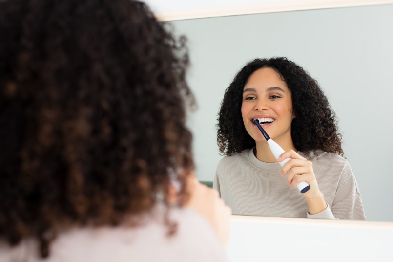 Braunhaarige, junge Frau steht vor dem Spiegel im Badezimmer, lächelt ihr Spiegelbild an und putzt dabei ihre Zähne mit der TB 15