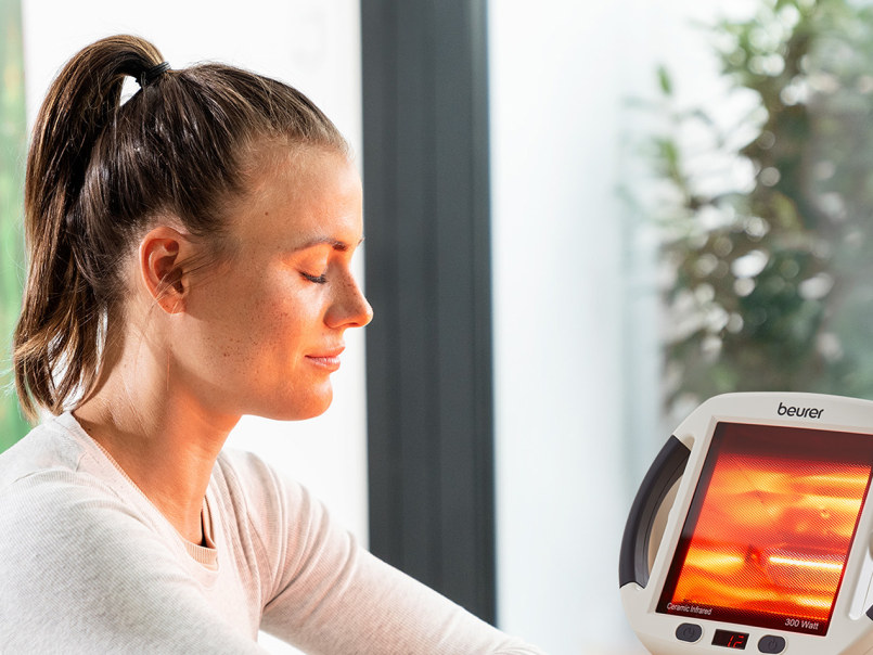 Eine Frau sitzt seitlich mit geschlossenen Augen vor einem Beurer IL 50 Infrarot-Wärmestrahler mit leuchtendem Heizfeld.