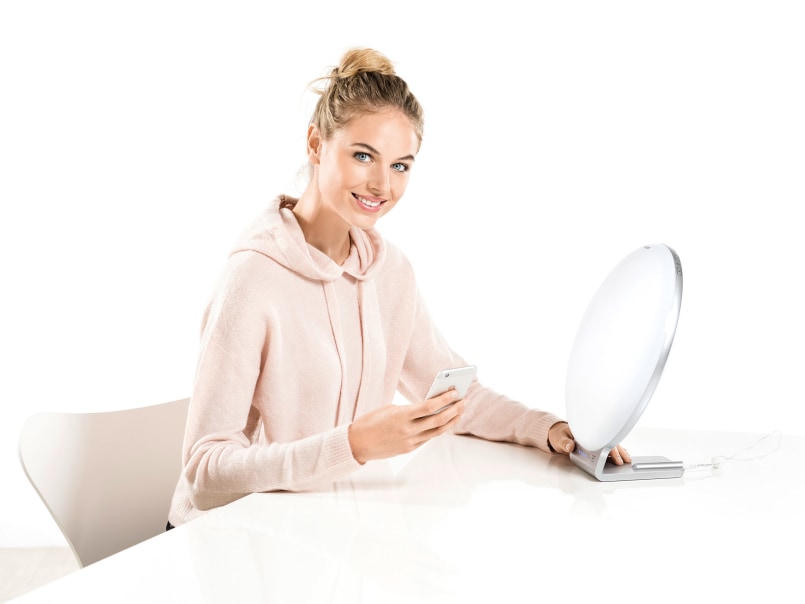 Eine Frau in hellem Kapuzenpullover sitzt an weißem Tisch und hält Smartphone, neben ihr steht die TL 100 Tageslichtlampe.