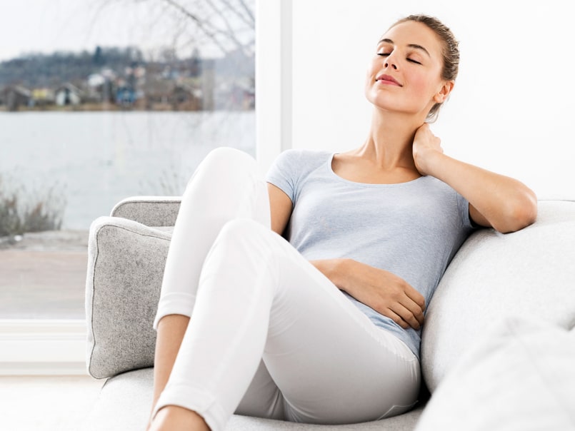 Frau in weißer Hose und grauem T-Shirt sitzt entspannt lächelnd auf einem hellen Sofa und hat die Hand an den Nacken gelegt.