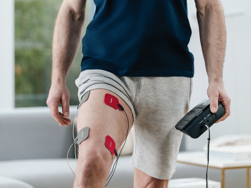 Mann trägt Shorts und hält EM 89 Heat Digital TENS/EMS-Gerät, am Oberschenkel sind vier Elektroden mit Kabeln befestigt.