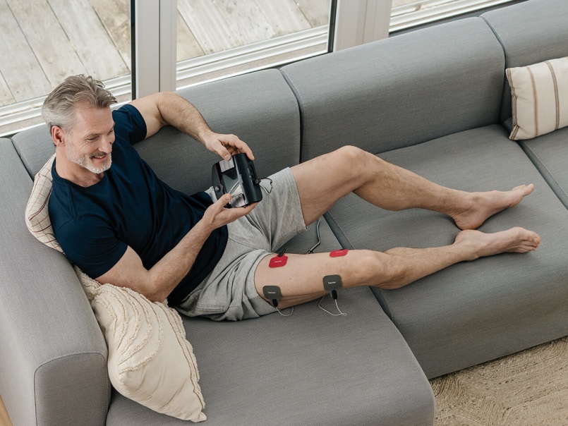 Mann liegt auf grauem Sofa, hält ein TENS/EMS-Gerät, an seinem ausgestreckten Oberschenkel sind vier Elektrodenpads befestigt.