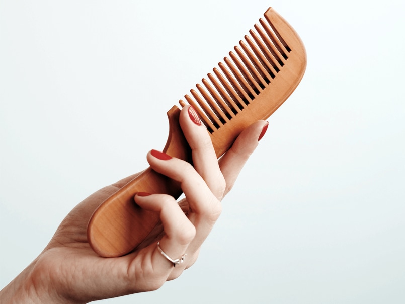 Hand mit rotem Nagellack hält einen braunen Holzkamm vor hellem Hintergrund.