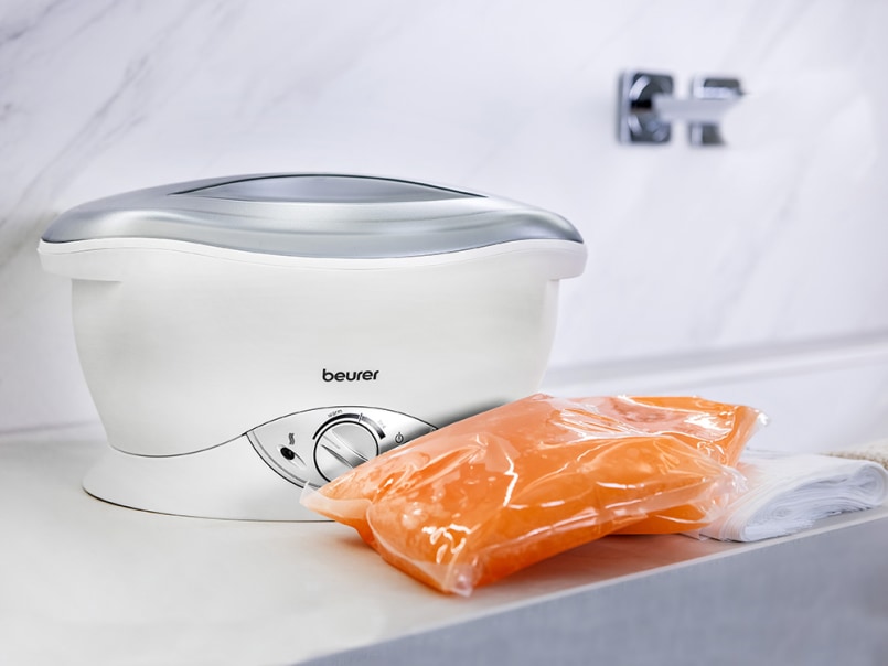 Beurer MP 70 Paraffinbad mit Temperaturregler, daneben orangefarbenes Paraffinwachs in Folienverpackung und ein weißes Tuch.