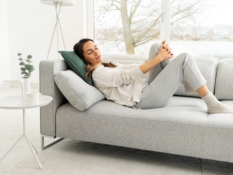 Frau liegt entspannt auf dem Sofa mit dem HK 48 Cosy Grey Sofa Heizkissen und lächelt mit geschlossenen Augen.