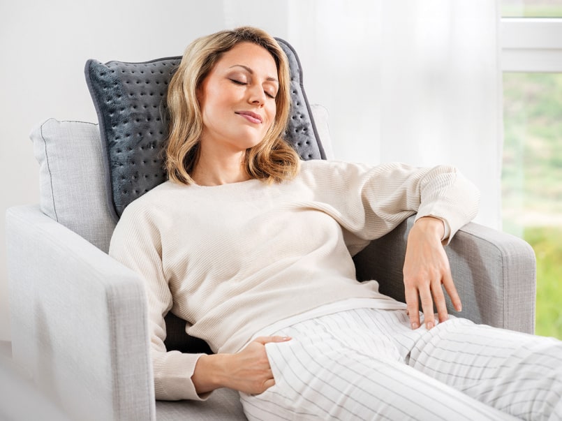 Person sitzt entspannt auf einem hellgrauen Sofa mit dem Beurer Heizkissen im Nacken.