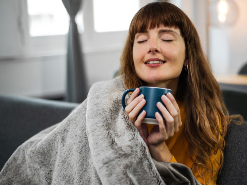 Person mit langen Haaren sitzt lächelnd auf Sofa, hält eine Tasse mit beiden Händen und ist mit der Wärmezudecke bedeckt.