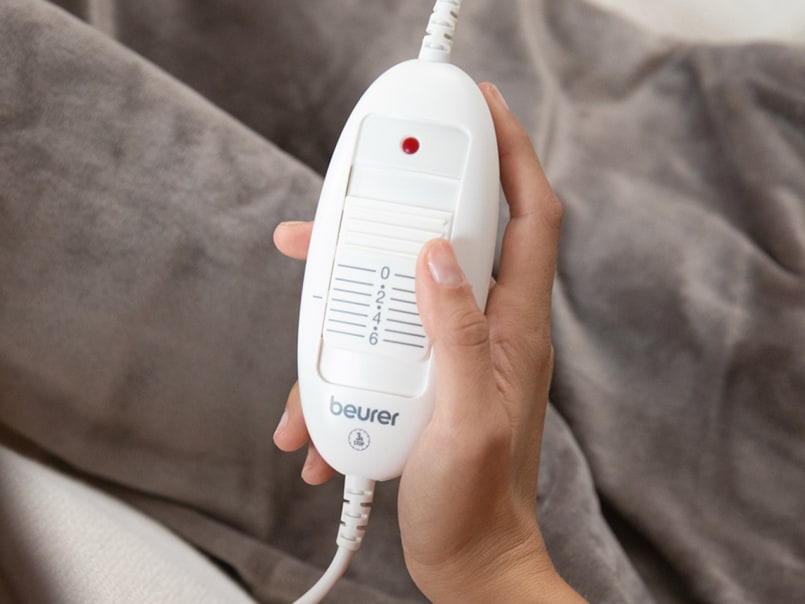 Hand hält weißen Beurer-Regler mit Schiebeschalter und roter Kontrollleuchte vor grauem Stoffhintergrund.