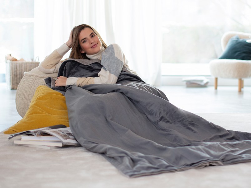 Person liegt entspannt auf dem Boden, eingehüllt in HD 75 Wärmezudecke, neben ihr aufgeschlagene Zeitschriften und Kissen.