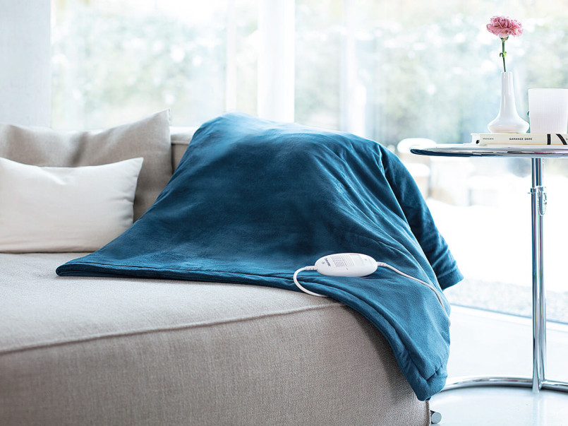 Blaue HD 75 Wärmezudecke liegt auf einem hellen Sofa neben einem runden Beistelltisch mit einer Vase und einem Buch.