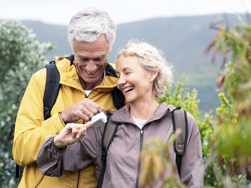 Älteres Paar im Outdoor-Outfit, die Frau hält ein weißes Pulsoximeter, der Mann zeigt darauf.
