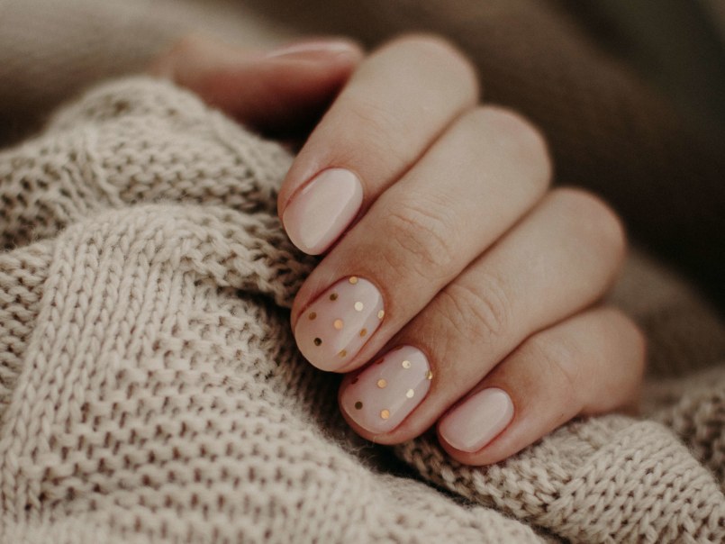 Hand mit gepflegten, kurz geschnittenen Nägeln, zwei Nägel mit goldfarbenen Punkten, auf grob gestricktem Stoff liegend.