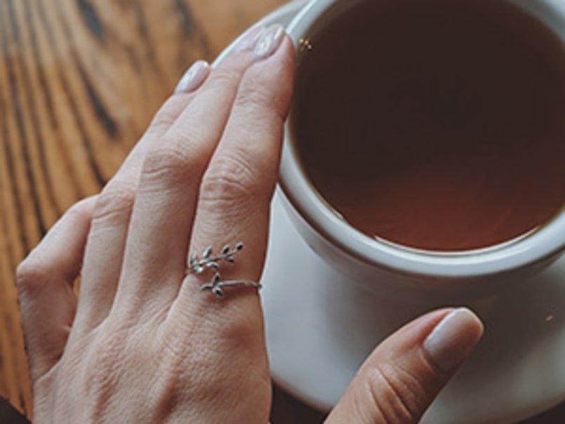 Hand mit gepflegten Nägeln und silbernem Ring mit Blattmotiv hält eine Tasse Kaffee auf einem Holztisch.