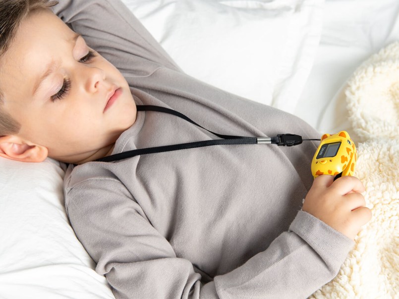 Kind liegt im Bett und benutzt gelbes Pulsoximeter mit Giraffenmuster an einem schwarzen Band.