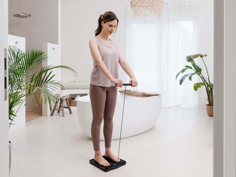 Sportlich gekleidete, junge Frau in einem modernen Badezimmer benutzt die BF 990 Diagnosewaage mit ausgezogenem Handteil
