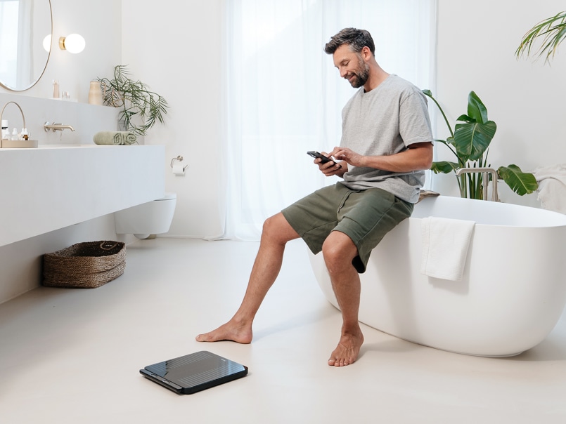 Ein junger Mann sitzt in einem modernen Badezimmer auf dem Rand der Badewanne und hat sein Smartphone in der Hand