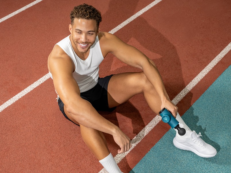2 Männer in Sport outfits sitzen auf einem blauen Boden und der linke hält die Massage Gun in der Hand