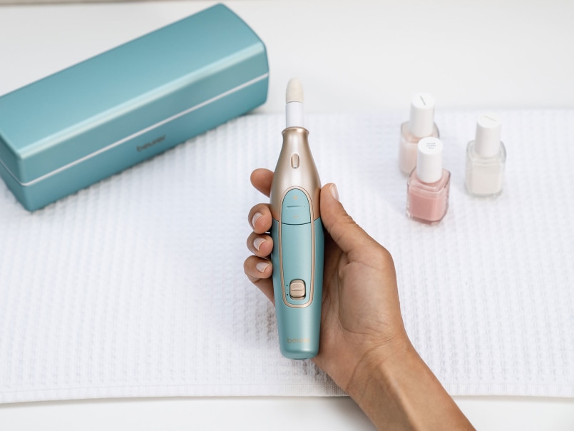 Der Beurer MP 84 Nagelfräser in mint liegt in der Hand und wird mit verschiedenen Nagellacken und der rechteckigen Ladebox präsentiert.