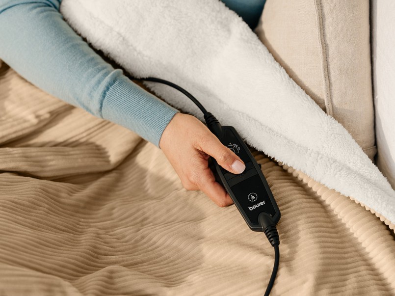 Hand hält schwarzen Beurer-Regler auf einer beige-weißen Wärmezudecke