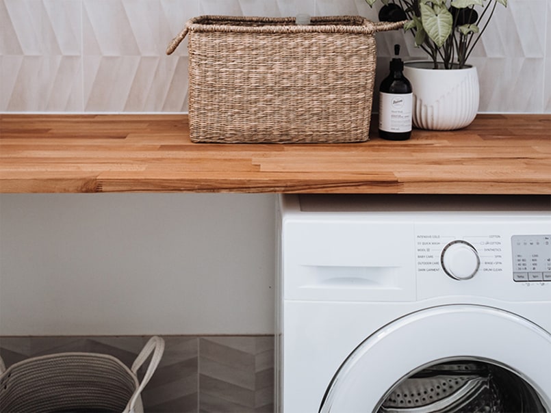 Waschmaschine unter einer Holzarbeitsplatte, darauf ein Korb, eine schwarze Flasche und eine Pflanze in einem weißen Topf