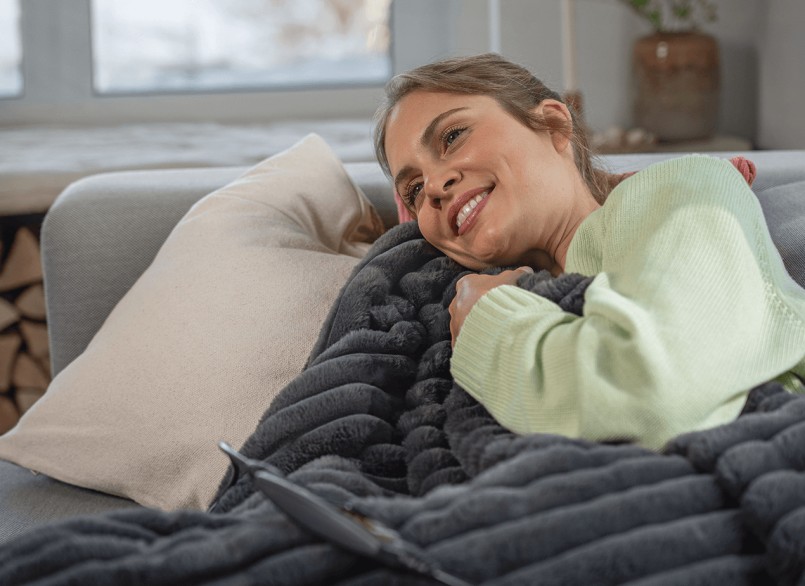Junge Frau mit grünem Pulli liegt eingekuschelt in der grauen Beurer Heizdecke Stockholm auf einem Sofa