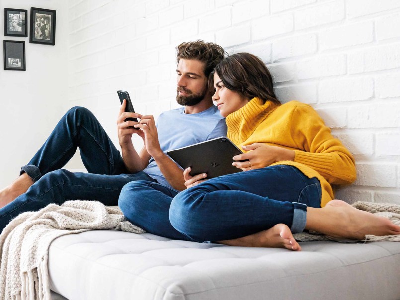 Frau und Mann sitzen mit Smartphone und Tablet in der Hand auf dem Sofa
