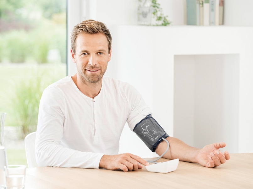 Mann sitzt an einem Tisch mit einem am Oberarm angelegten Blutdruckmessgerät und hält das Messgerät in der Hand