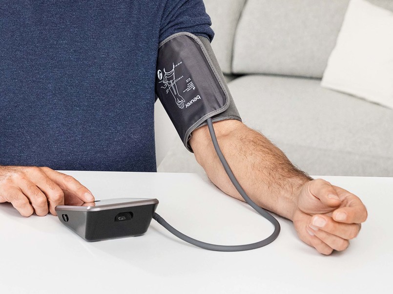 Mann sitzt am Tisch und misst Blutdruck am Oberarm mit dem Beurer BM 64, drückt mit Finger auf den Startknopf des Geräts