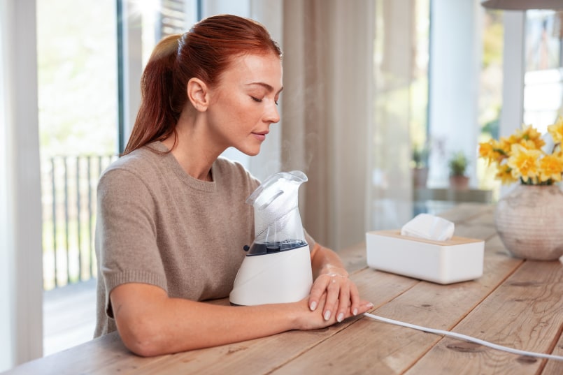 Frau sitzt am Tisch, Beurer SI 31 vor ihr zwischen ihren Armen, sie atmet Dampf durch Nase ein und hat Augen geschlossen