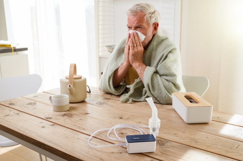 Älterer Mann sitzt am Tisch und putzt sich die Nase, er hat eine Decke um die Schultern, vor ihm steht eine Teekanne und Tee, eine Taschentücherbox und der Beuer IH 15 Inhalator